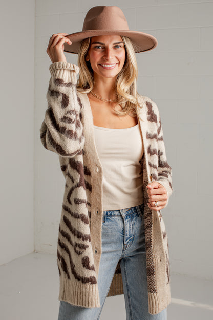 A woman wearing a beige and brown leopard-print knit cardigan, a brown wide-brim hat, a cream tank top, and light blue jeans, standing against a light gray wall.
