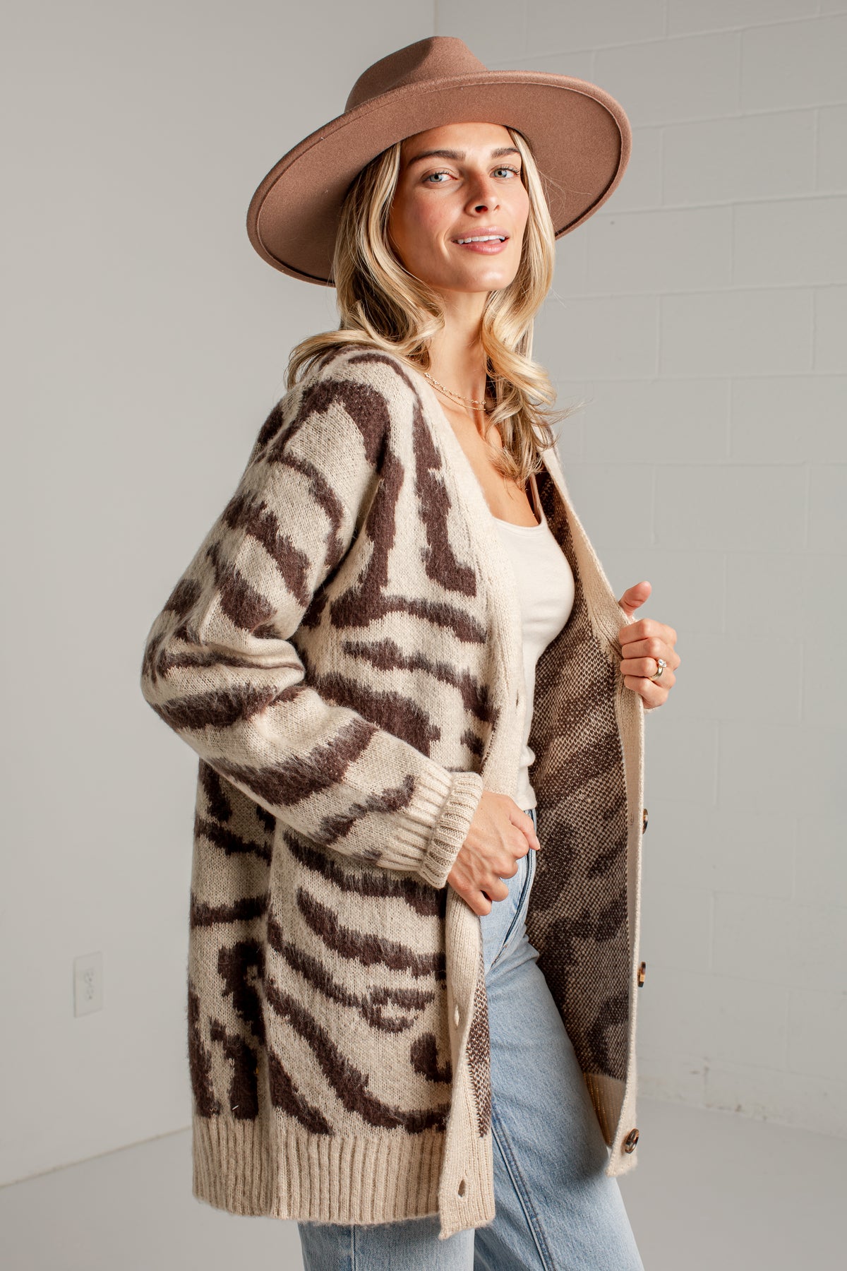 A woman wearing a beige and brown zebra-print knit cardigan, a brown wide-brim hat, a white top, and light blue jeans, standing against a light gray brick wall.