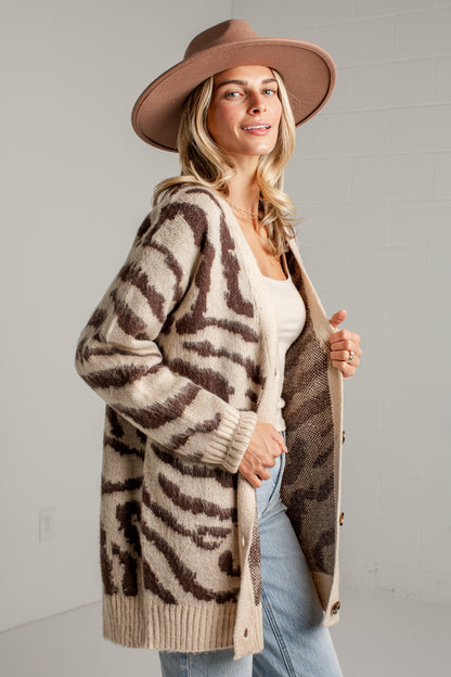 A woman wearing a beige and brown zebra-print knit cardigan, a brown wide-brim hat, a white top, and light blue jeans, standing against a light gray brick wall.