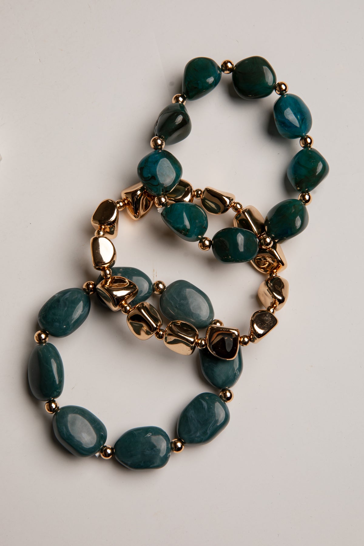 A beaded stretch bracelet featuring dark teal-green polished stones and gold-toned metal links, arranged in a loop on a light gray background.