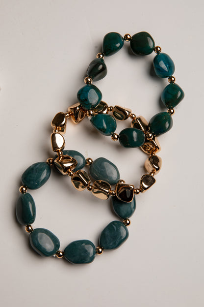A beaded stretch bracelet featuring dark teal-green polished stones and gold-toned metal links, arranged in a loop on a light gray background.