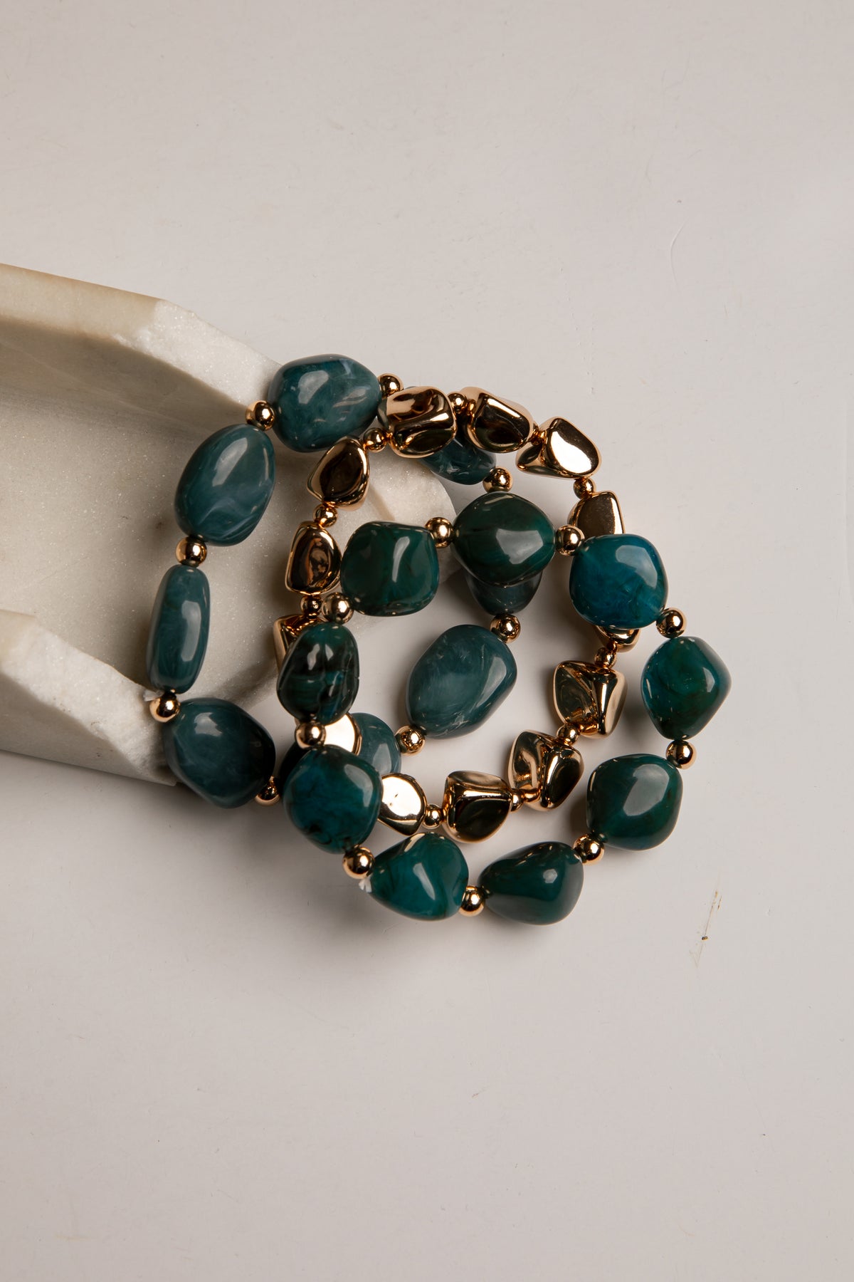 A multi-strand statement necklace featuring polished dark teal stone beads and antique gold-toned metal links, displayed on a cream ceramic stand against a light gray background.