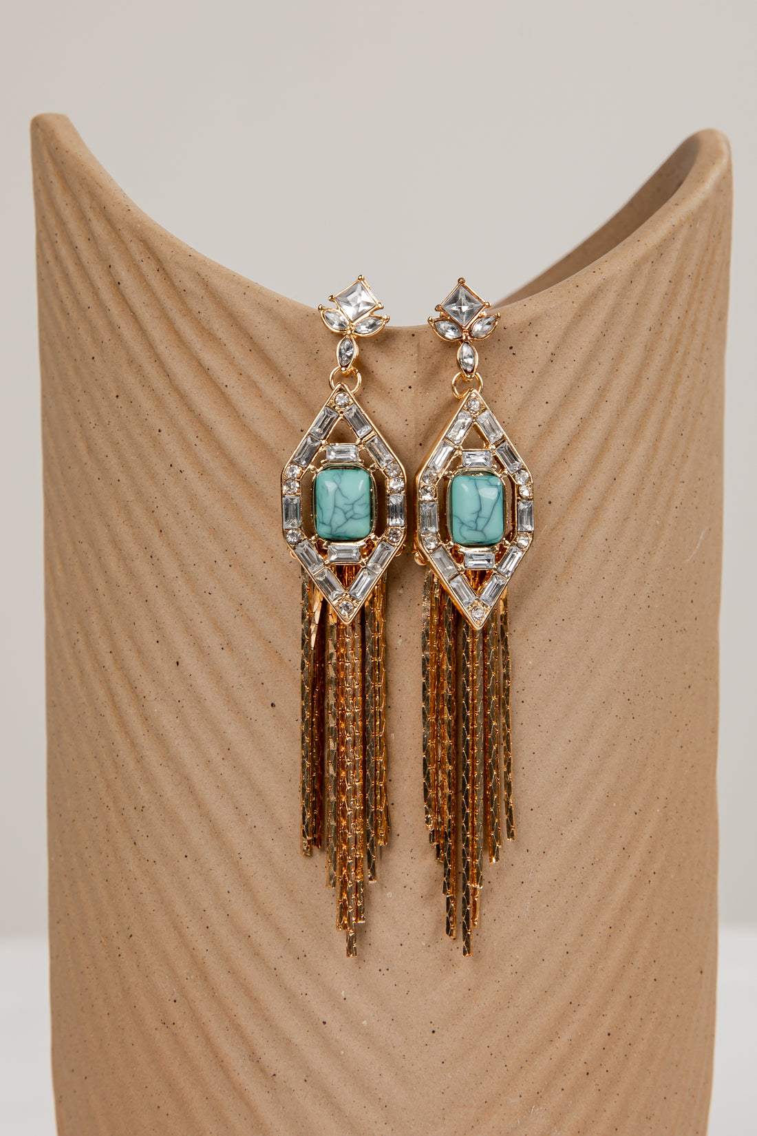 Pair of earrings with turquoise stones and gold fringes on a beige stand.