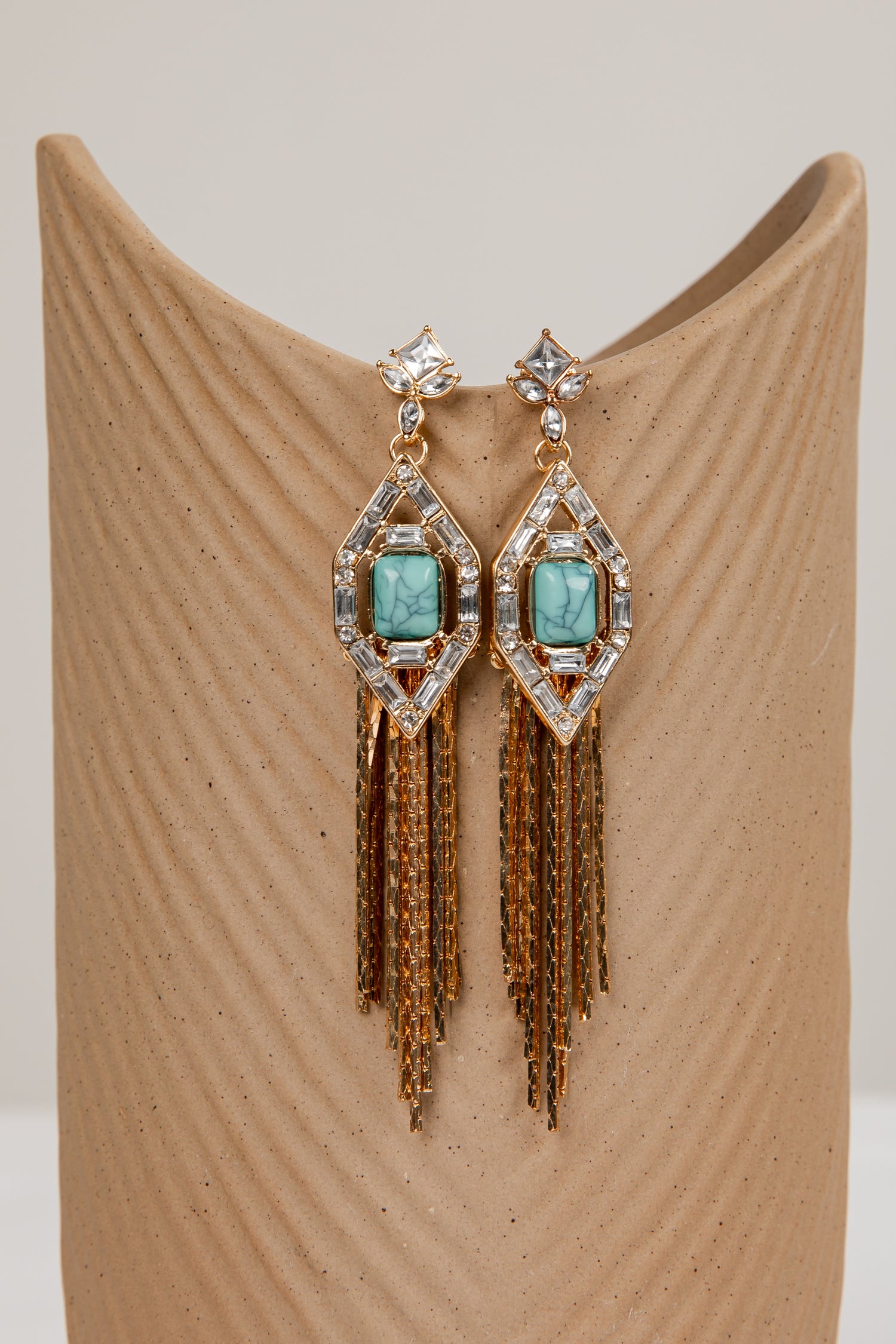 Pair of earrings with turquoise stones and gold fringes on a beige stand.