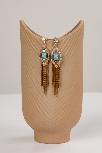 Pair of earrings with turquoise stones and gold fringes on a beige stand.