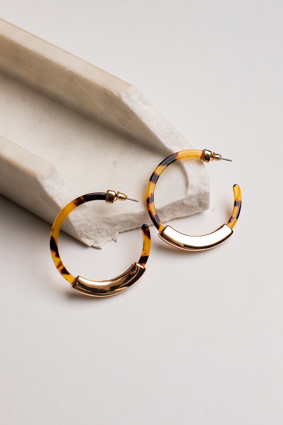 Tortoiseshell hoop earrings with gold-toned metal accents, displayed on a light gray surface next to a white stone prop.