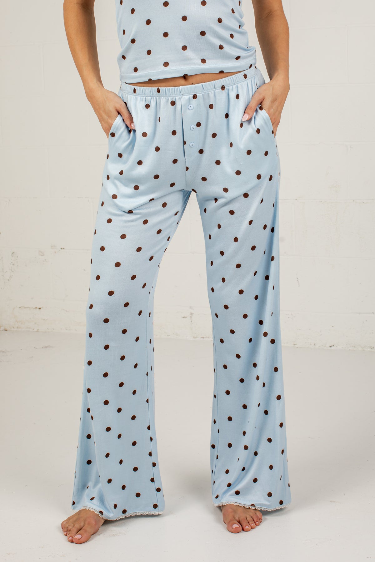 A person wearing light blue pajama pants with a brown polka-dot pattern, standing barefoot against a white wall.