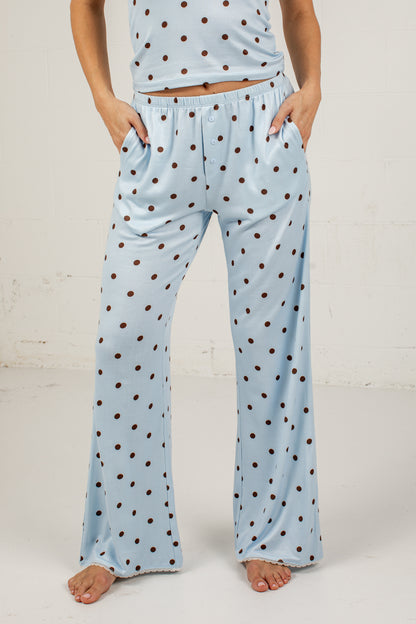 A person wearing light blue pajama pants with a brown polka-dot pattern, standing barefoot against a white wall.