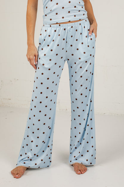 A person wearing light blue wide-leg pajama pants with a brown polka dot pattern, shown from the waist down against a white studio wall.