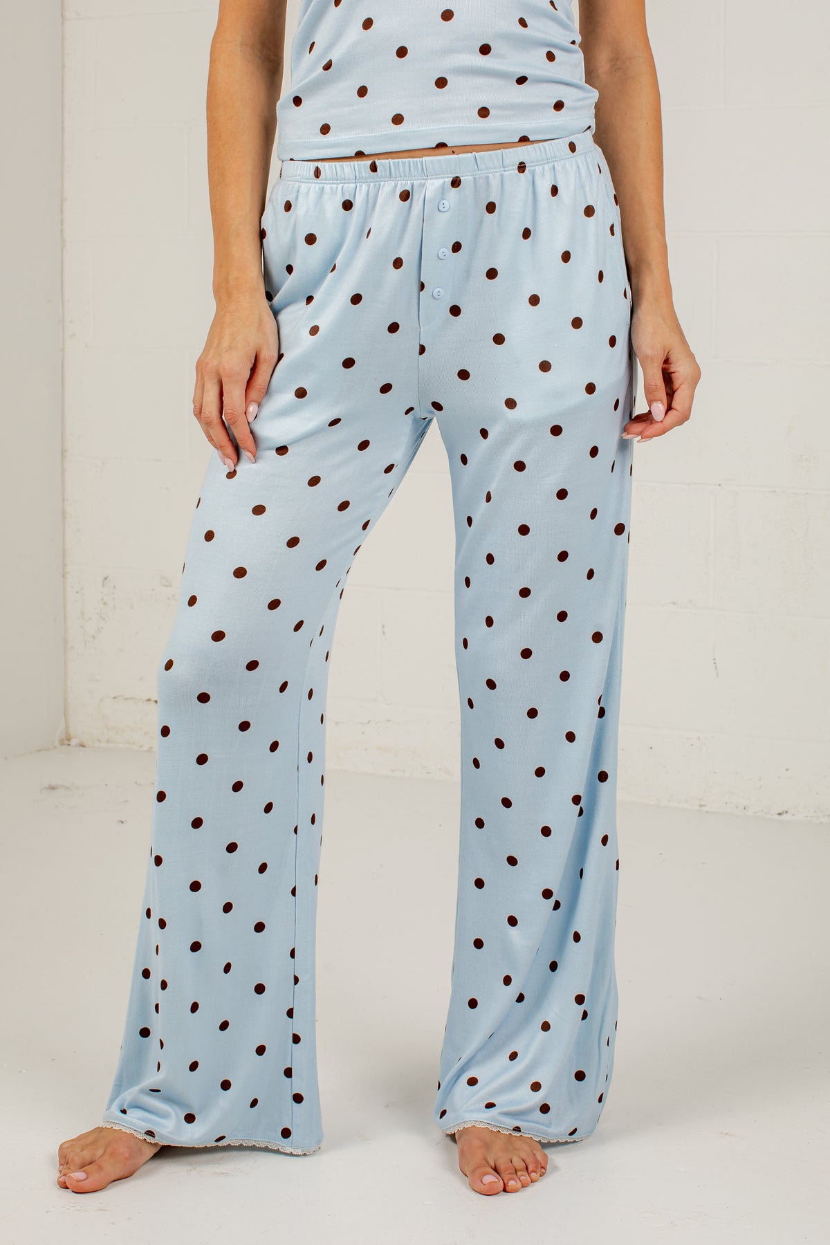 A person wearing light blue pajama pants with an allover brown polka dot pattern, shown from the waist down against a plain off-white wall.