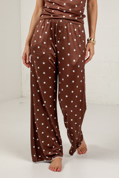 A person wearing brown wide-leg pajama pants with a white polka dot pattern, shown from the waist down in a studio setting.