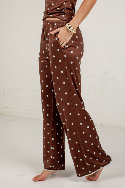A person wearing brown wide-leg pajama pants with white polka dots, standing barefoot against a white wall.