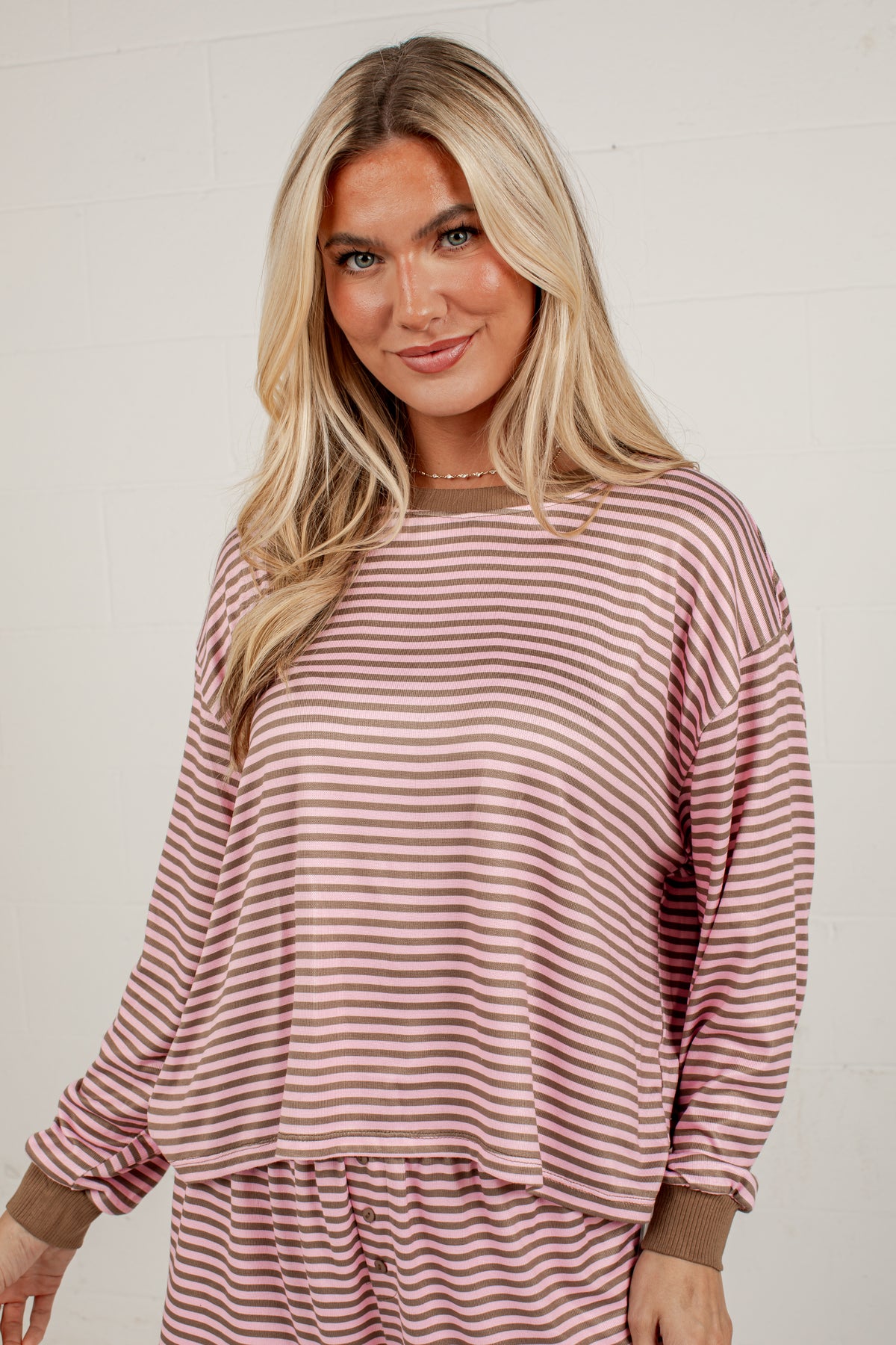 A woman modeling a long-sleeve dusty rose and brown striped lounge top against a light off-white brick wall.