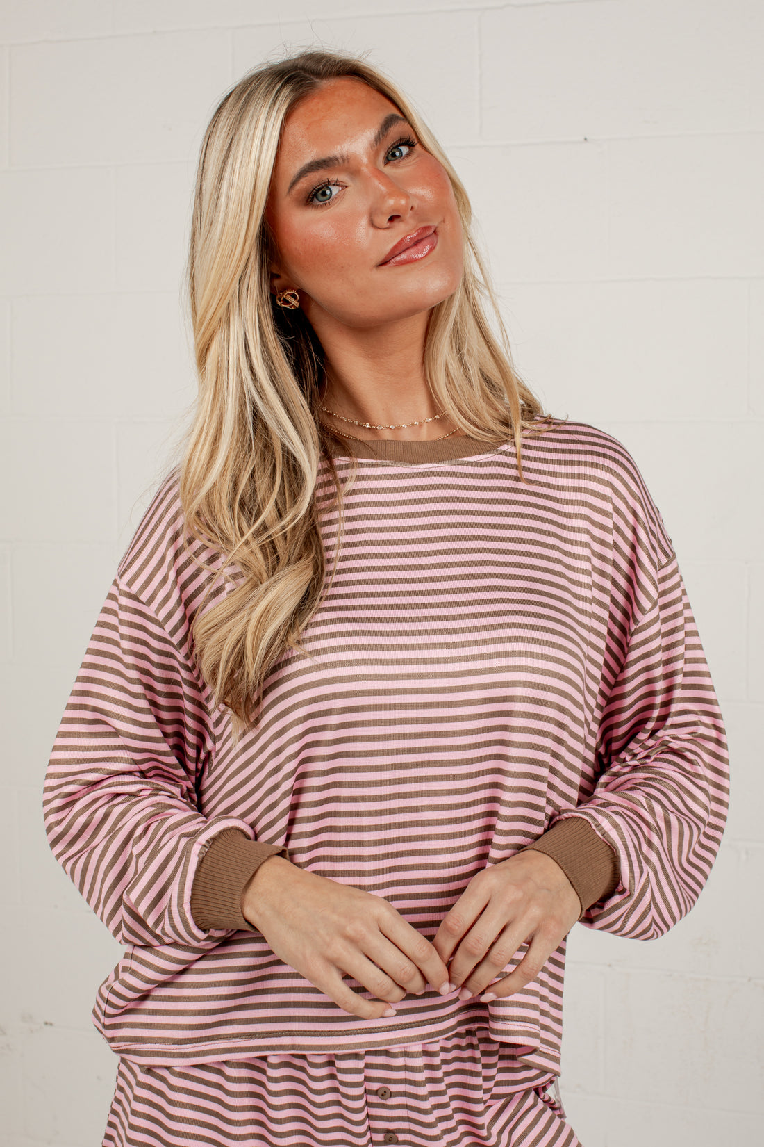 A blonde woman wearing a long-sleeve crewneck top with horizontal dusty rose and brown stripes, paired with matching striped pants, standing against a white brick wall.