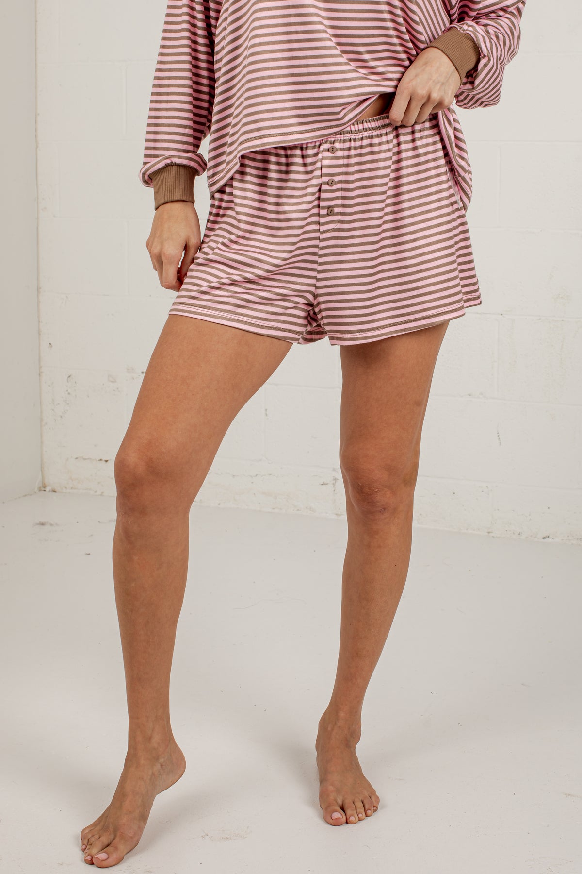 A model wearing a matching two-piece pajama set with a pink and white striped long-sleeve top and shorts, standing barefoot against a white brick wall.