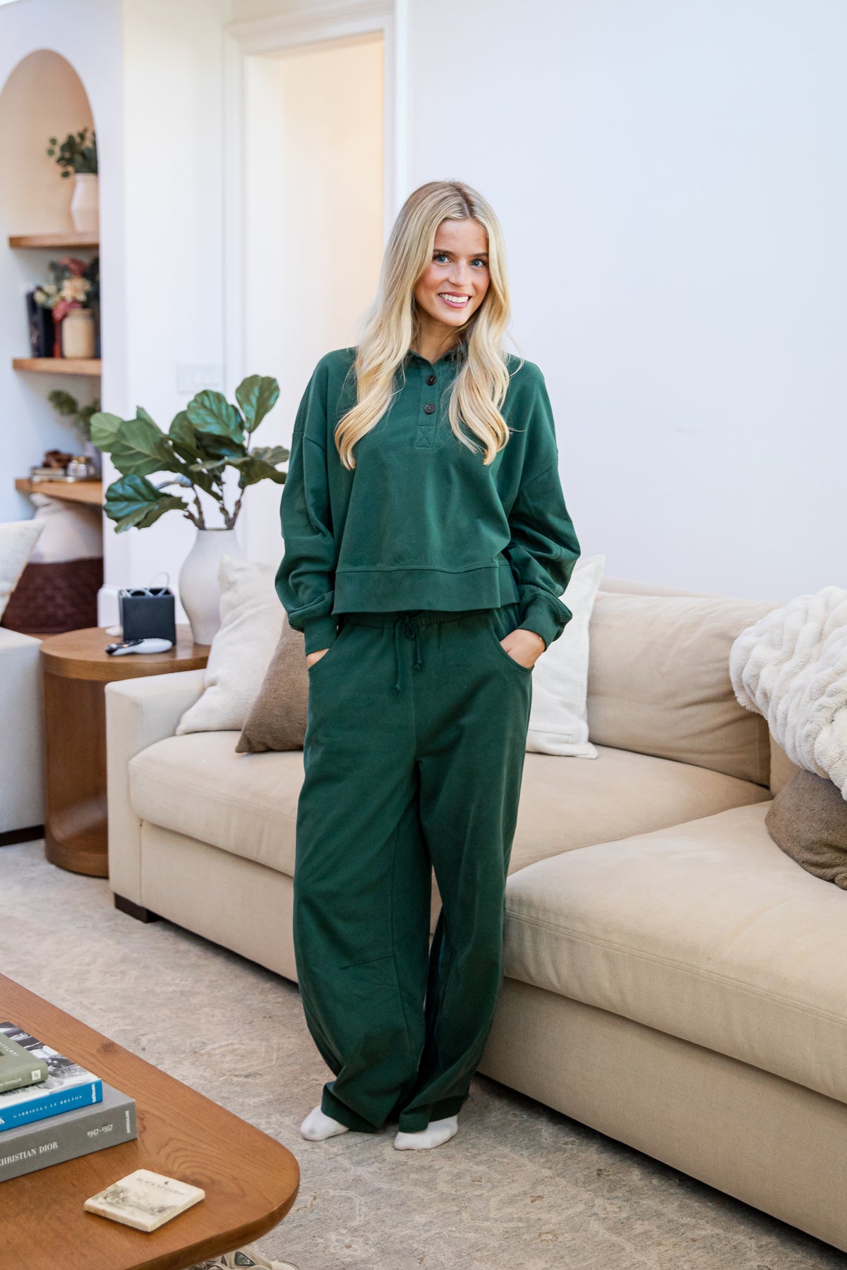 A woman stands in a living room wearing a matching dark green sweatshirt and wide-leg sweatpants set. The room features a beige sofa, a wooden coffee table, and a potted plant in the background.