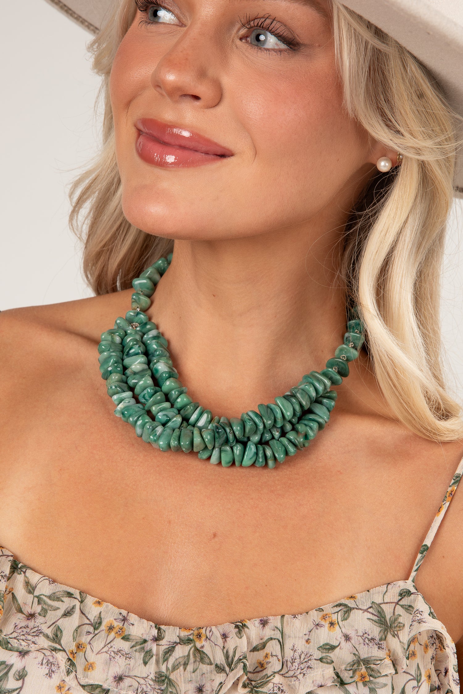 A close-up of a woman wearing a chunky, multi-strand turquoise green stone necklace, styled with a floral top and a light-colored hat.