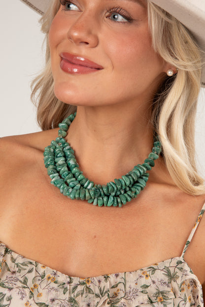 A close-up of a woman wearing a chunky, multi-strand turquoise green stone necklace, styled with a floral top and a light-colored hat.