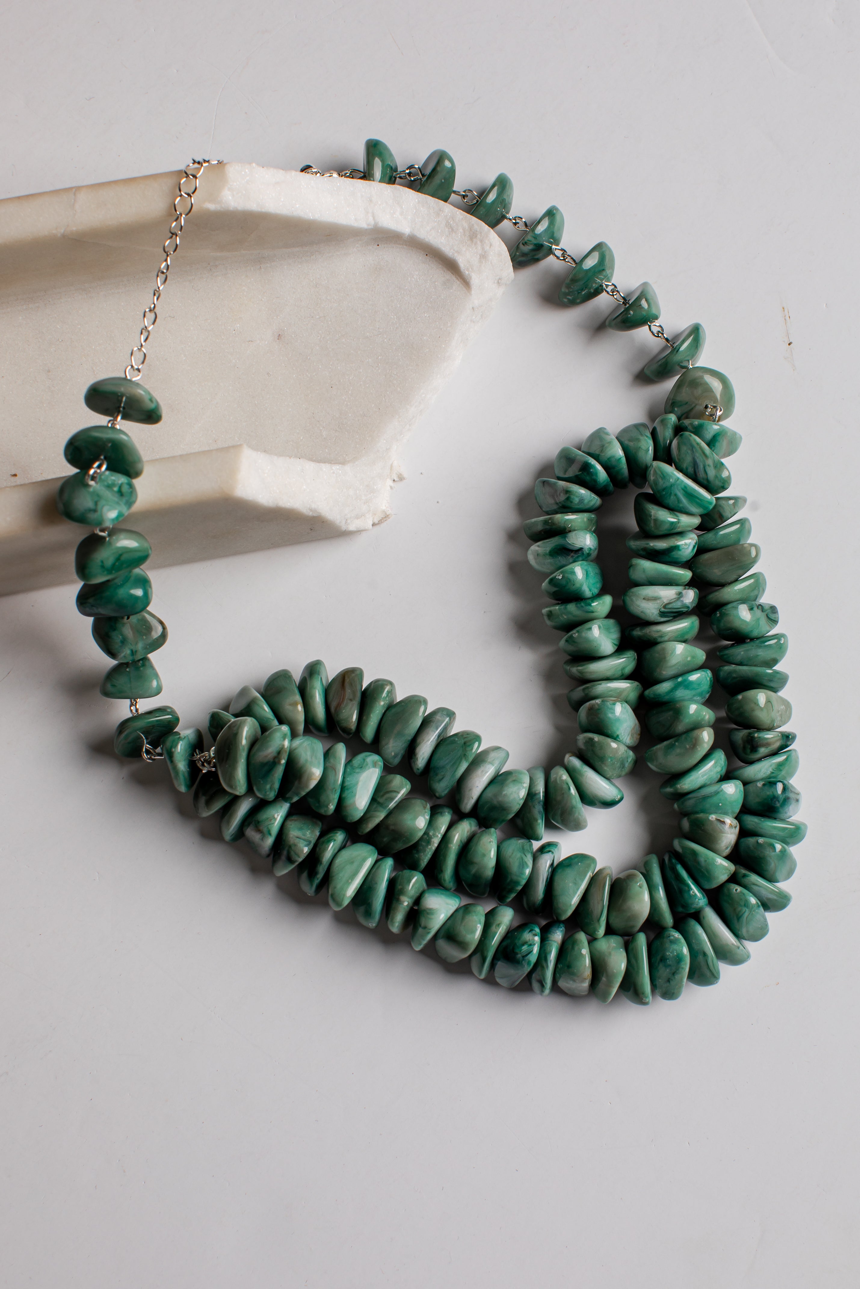 A multi-strand necklace made of polished green stone chips, draped over a light gray surface next to a white ceramic dish. The necklace features silver-toned metal chain links and a clasp.