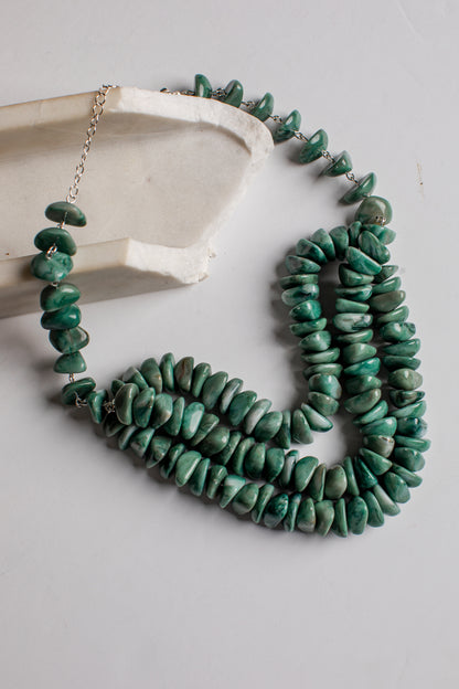 A multi-strand necklace made of polished green stone chips, draped over a light gray surface next to a white ceramic dish. The necklace features silver-toned metal chain links and a clasp.