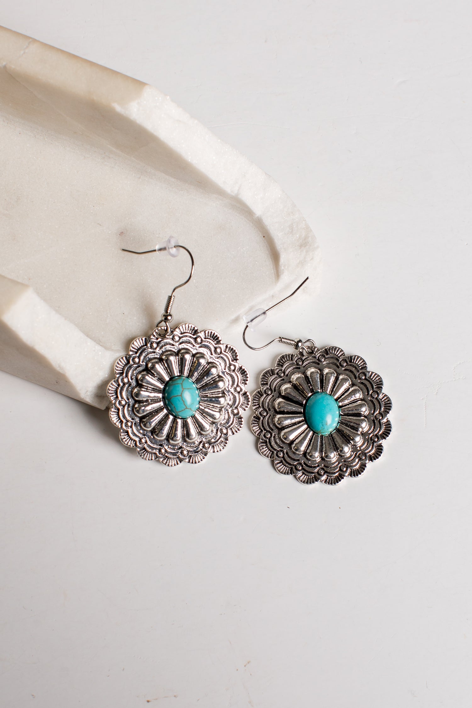A pair of silver-toned dangle earrings with intricate floral filigree detailing and a central turquoise stone, displayed on a light cream textured surface.