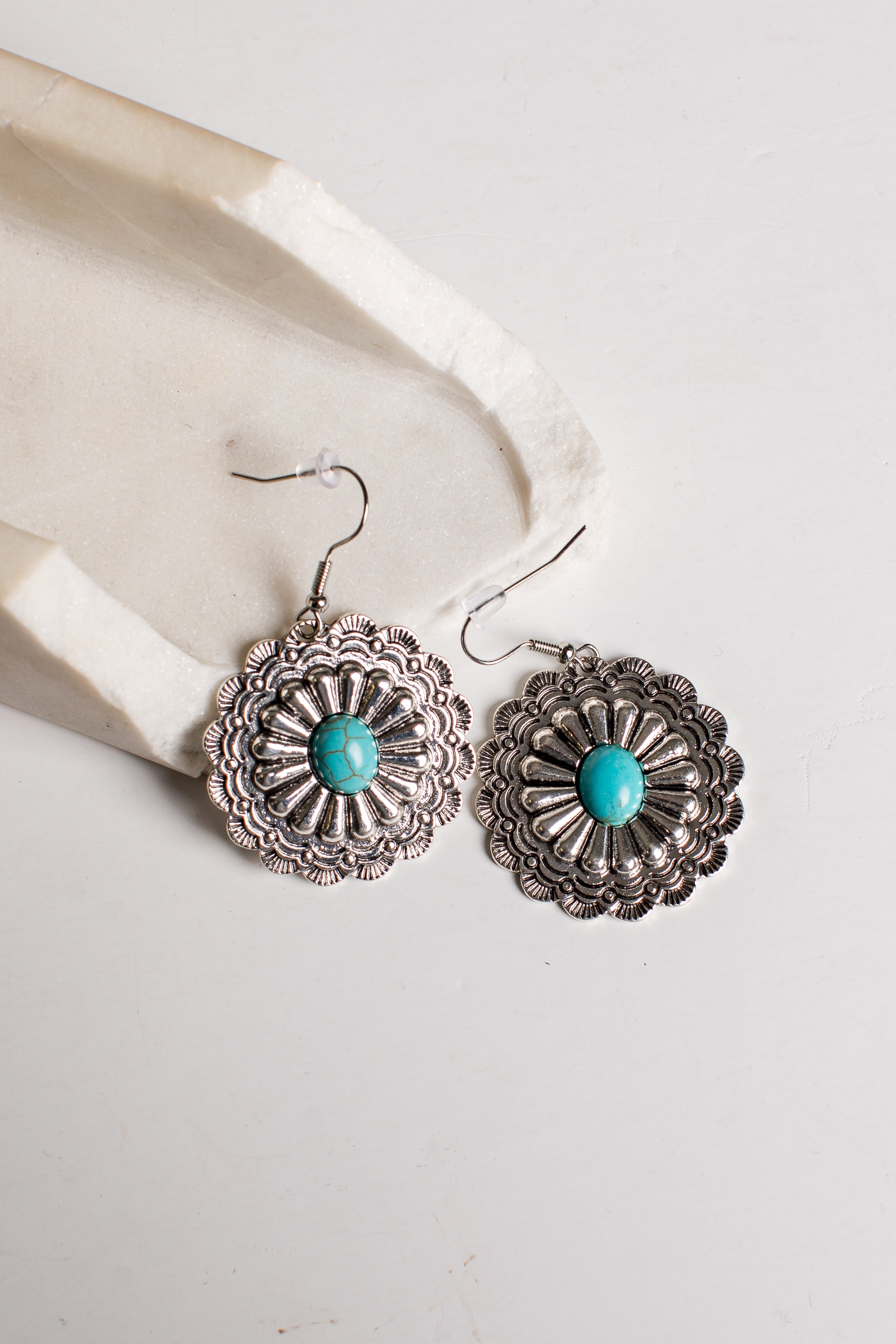A pair of silver-toned dangle earrings with intricate floral filigree detailing and a central turquoise stone, displayed on a light cream textured surface.