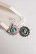 A pair of silver-toned dangle earrings with intricate floral filigree detailing and a central turquoise stone, displayed on a light cream textured surface.