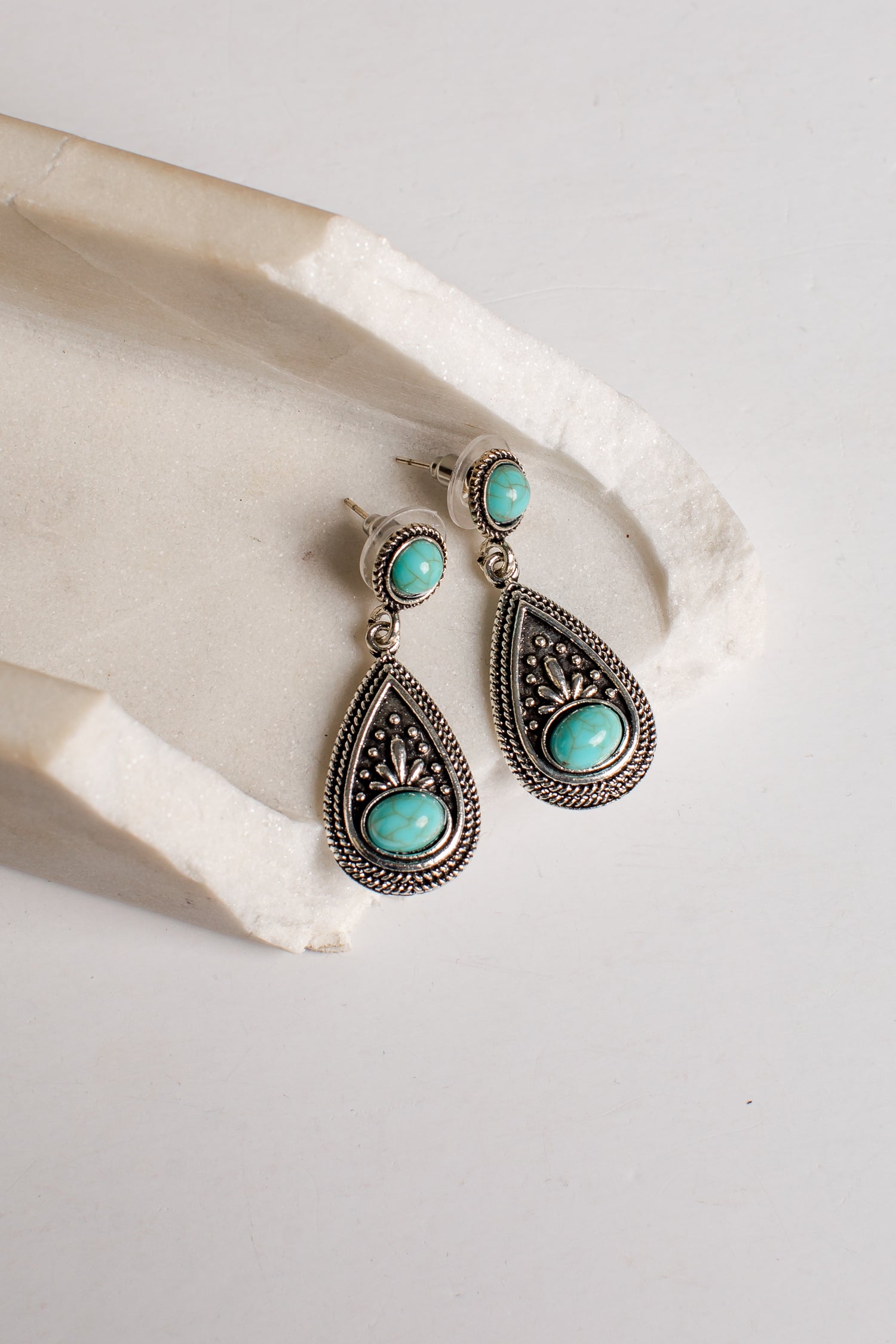 A pair of silver-toned teardrop dangle earrings with oval turquoise stones, displayed on a textured white stone prop against a light neutral background.