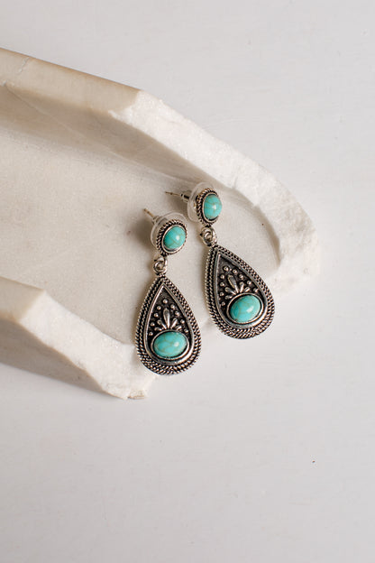 A pair of silver-toned teardrop dangle earrings with oval turquoise stones, displayed on a textured white stone prop against a light neutral background.