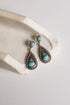 A pair of silver-toned teardrop dangle earrings with oval turquoise stones, displayed on a textured white stone prop against a light neutral background.