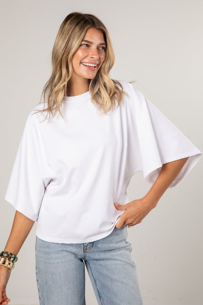 A woman wearing a white short-sleeve tunic top with wide, draped sleeves and light-wash blue jeans, standing against a plain light gray studio background.
