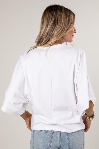A woman shown from the back wearing a white short-sleeve top with voluminous elbow-length sleeves and light blue jeans, against a plain light gray studio background.