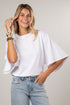 A woman wearing a white short-sleeve top with wide bell sleeves and light blue jeans, posing against a neutral light gray background.
