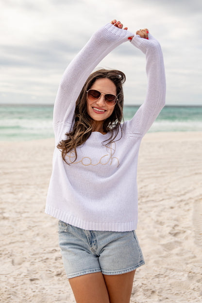 Woman on a beach wearing a white sweater with text and denim shorts, stretching her arms above her head.