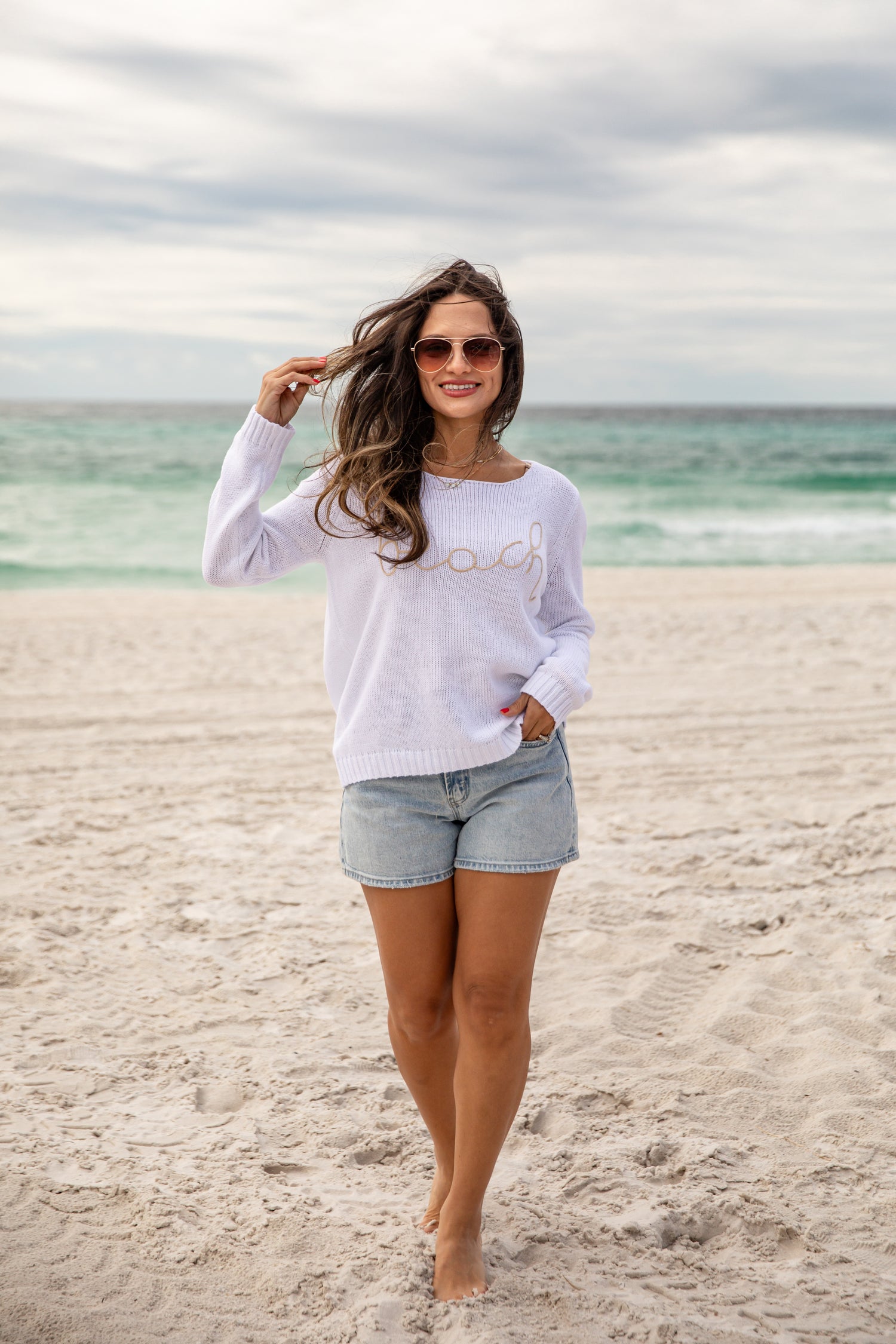 Woman standing on a beach wearing a white long-sleeve shirt and denim shorts.