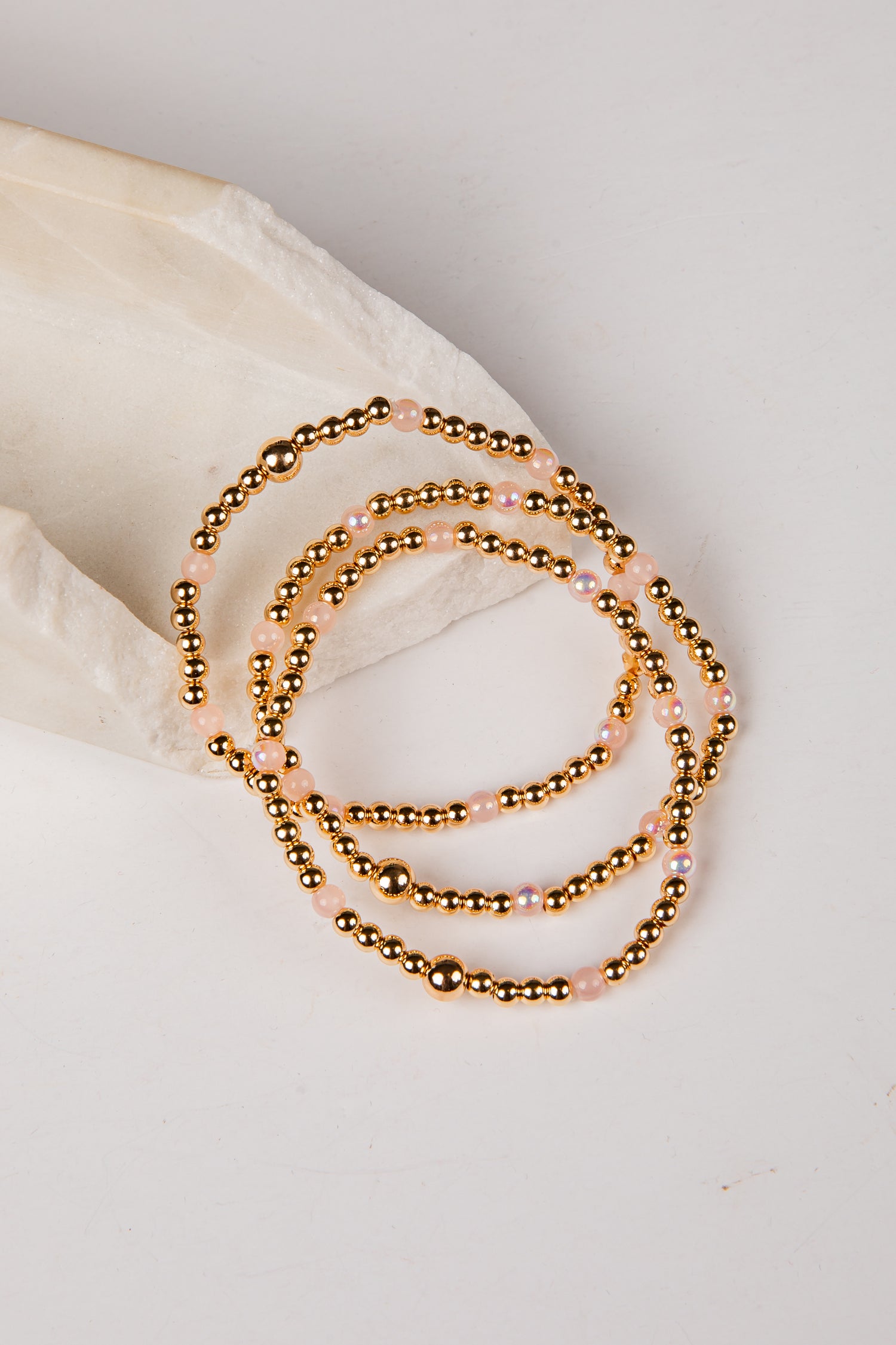 A stack of three beaded stretch bracelets with metallic gold beads and small iridescent pink beads, arranged on a light cream fabric prop against a white background.