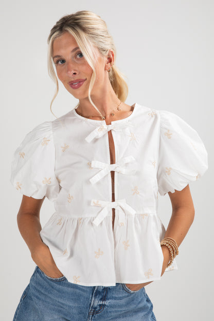 Woman wearing a white blouse with puff sleeves and floral patterns, standing against a plain background.