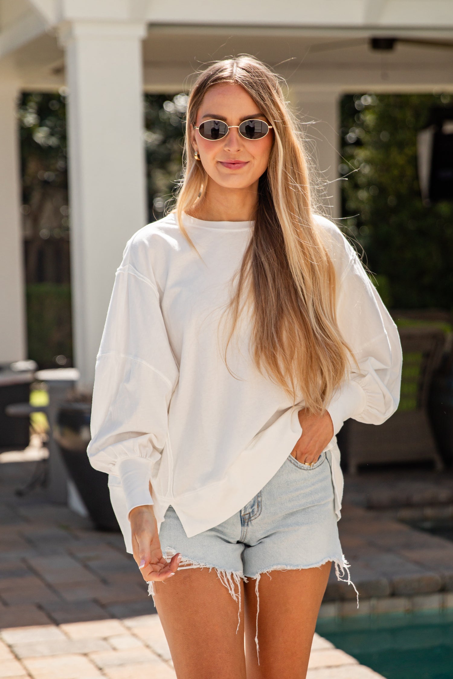 Woman wearing a white sweatshirt and denim shorts standing by a pool.