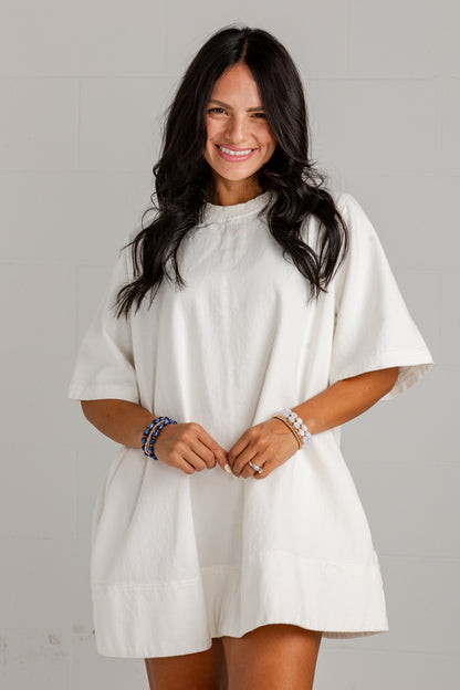 A woman with long dark hair wearing a loose-fitting, short-sleeved white linen dress or tunic with a gathered neckline, standing against a light gray brick wall.