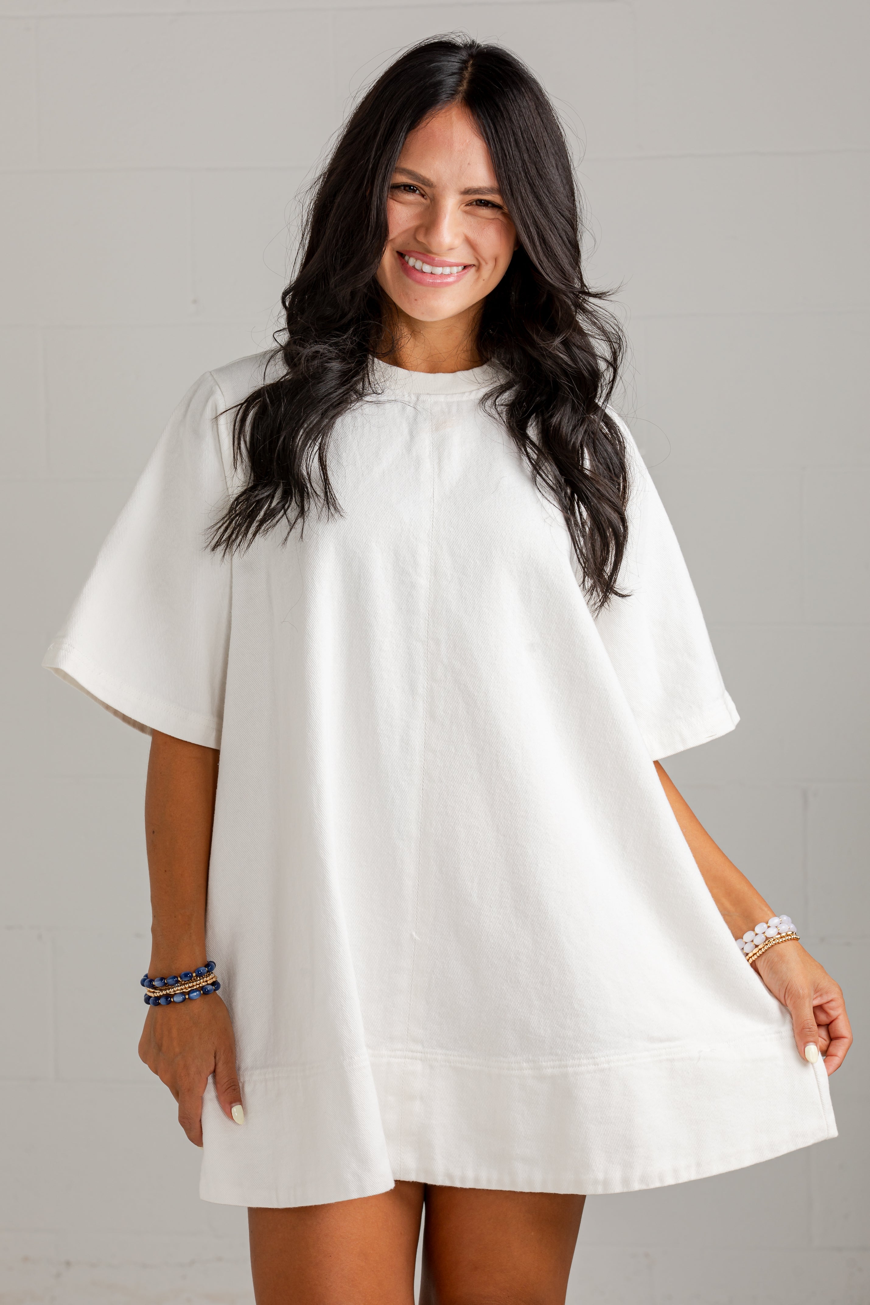 A woman modeling a loose, short-sleeve white tunic dress against a light gray wall.