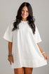 A woman modeling a loose, short-sleeve white tunic dress against a light gray wall.