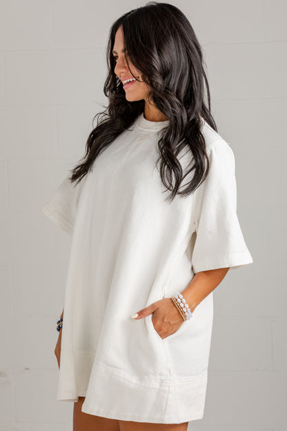A woman wearing an oversized white short-sleeve tunic top with side pockets, standing against a light gray wall.
