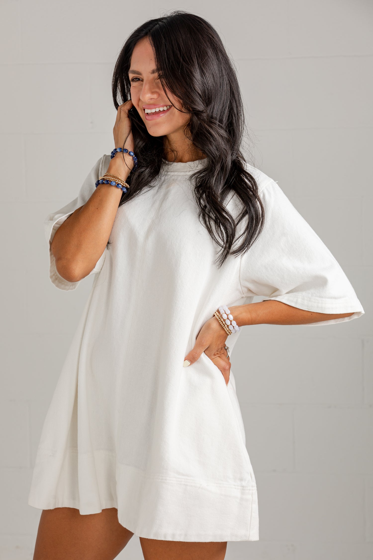 A woman wearing a loose white short-sleeve tunic dress with side pockets, posing against a light gray wall.