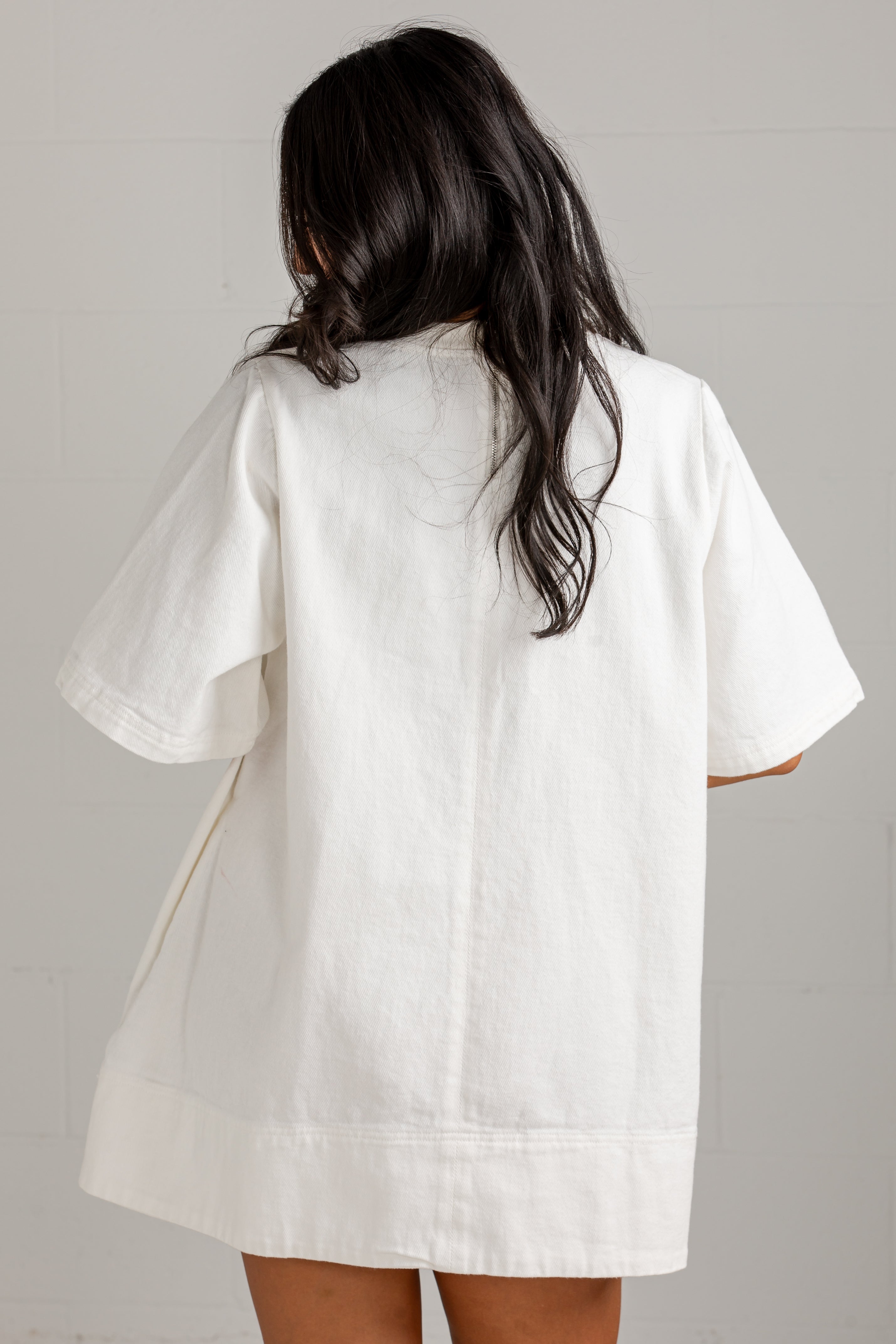 A woman viewed from behind wearing an oversized white short-sleeve shirt or tunic with a relaxed, loose fit and a slightly raw, casual hem.