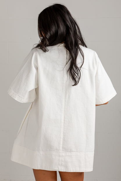 A woman viewed from behind wearing an oversized white short-sleeve shirt or tunic with a relaxed, loose fit and a slightly raw, casual hem.