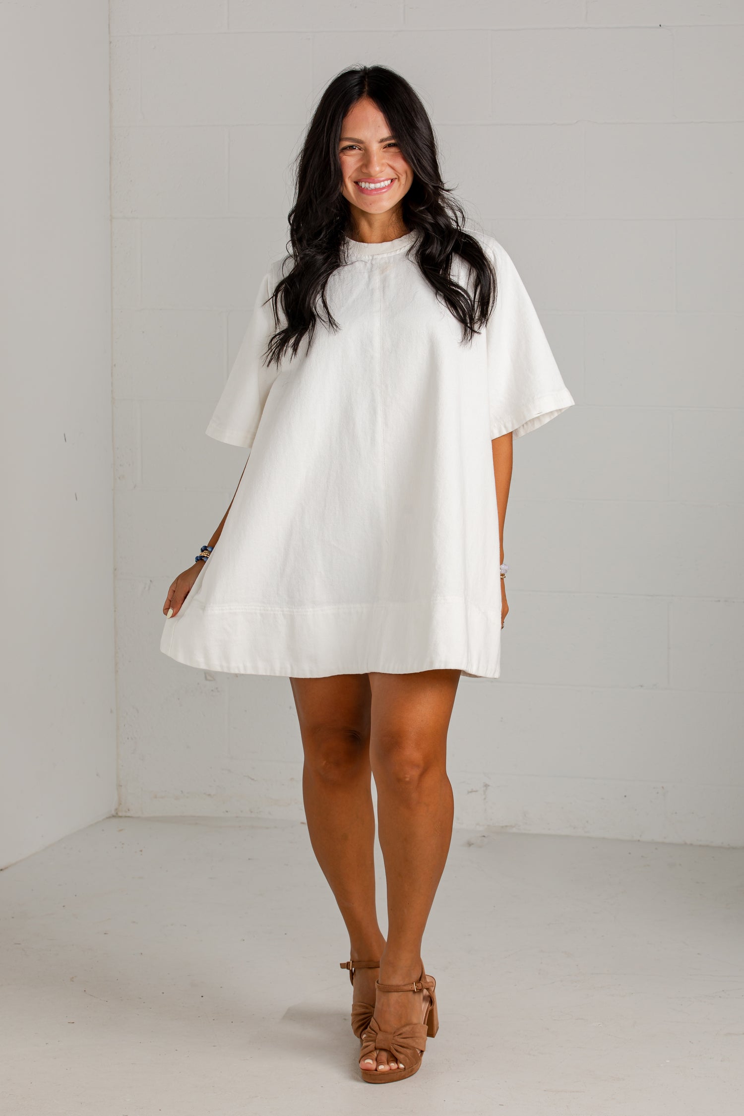 A woman modeling a white short-sleeve tunic dress with a high gathered neckline and a loose, flowing silhouette, paired with tan heeled sandals in a bright white studio setting.