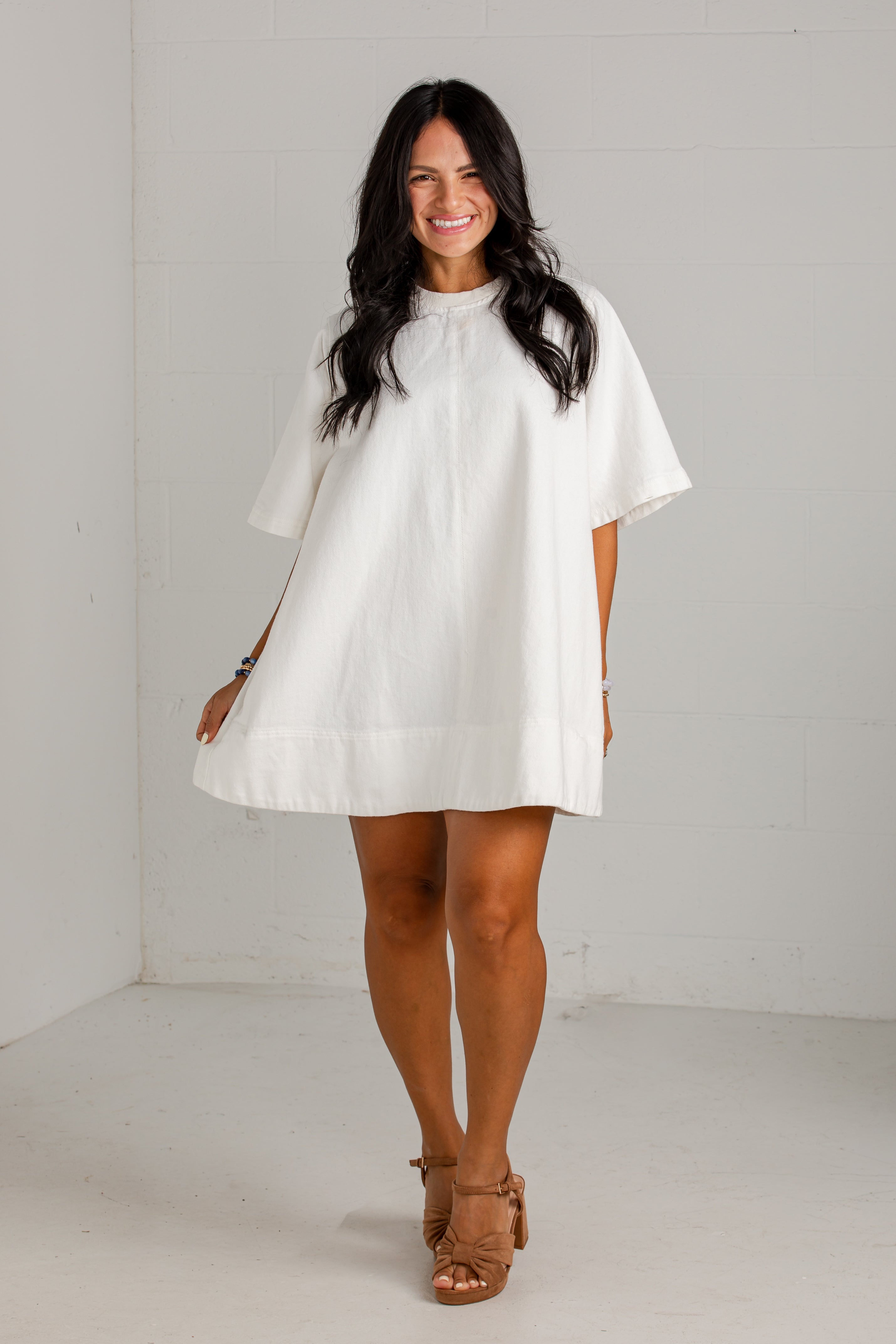 A woman modeling a white short-sleeve tunic dress with a high gathered neckline and a loose, flowing silhouette, paired with tan heeled sandals in a bright white studio setting.