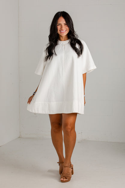 A woman modeling a white short-sleeve tunic dress with a high gathered neckline and a loose, flowing silhouette, paired with tan heeled sandals in a bright white studio setting.
