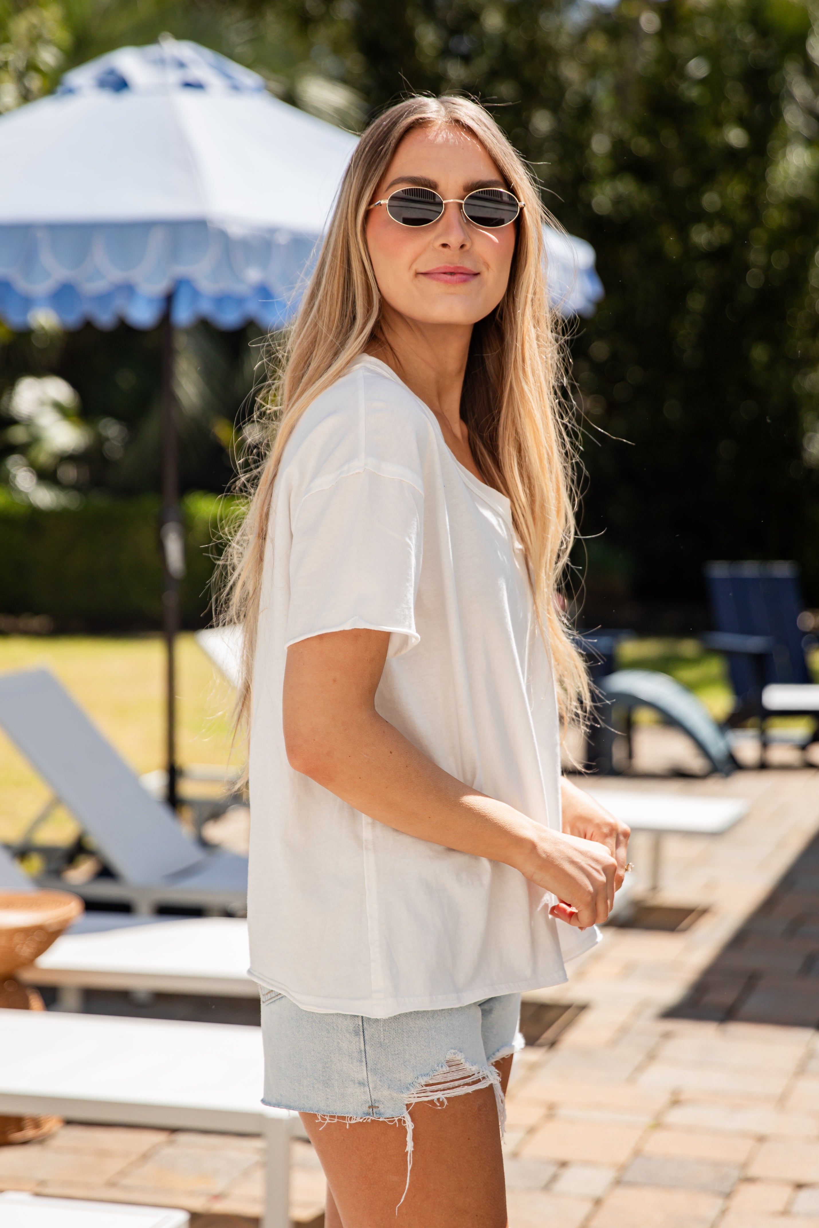 Woman in a white shirt and denim shorts standing by a poolside with sunglasses on.