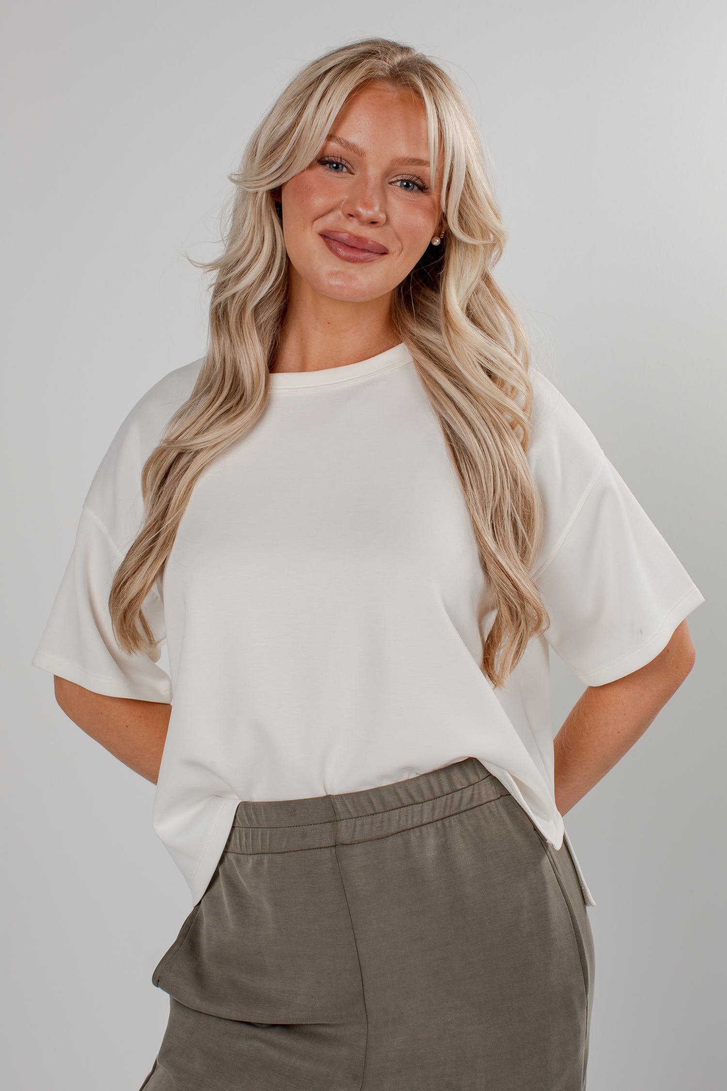 A blonde woman wearing a white short-sleeve crewneck top and gray pants, standing against a light gray studio background.